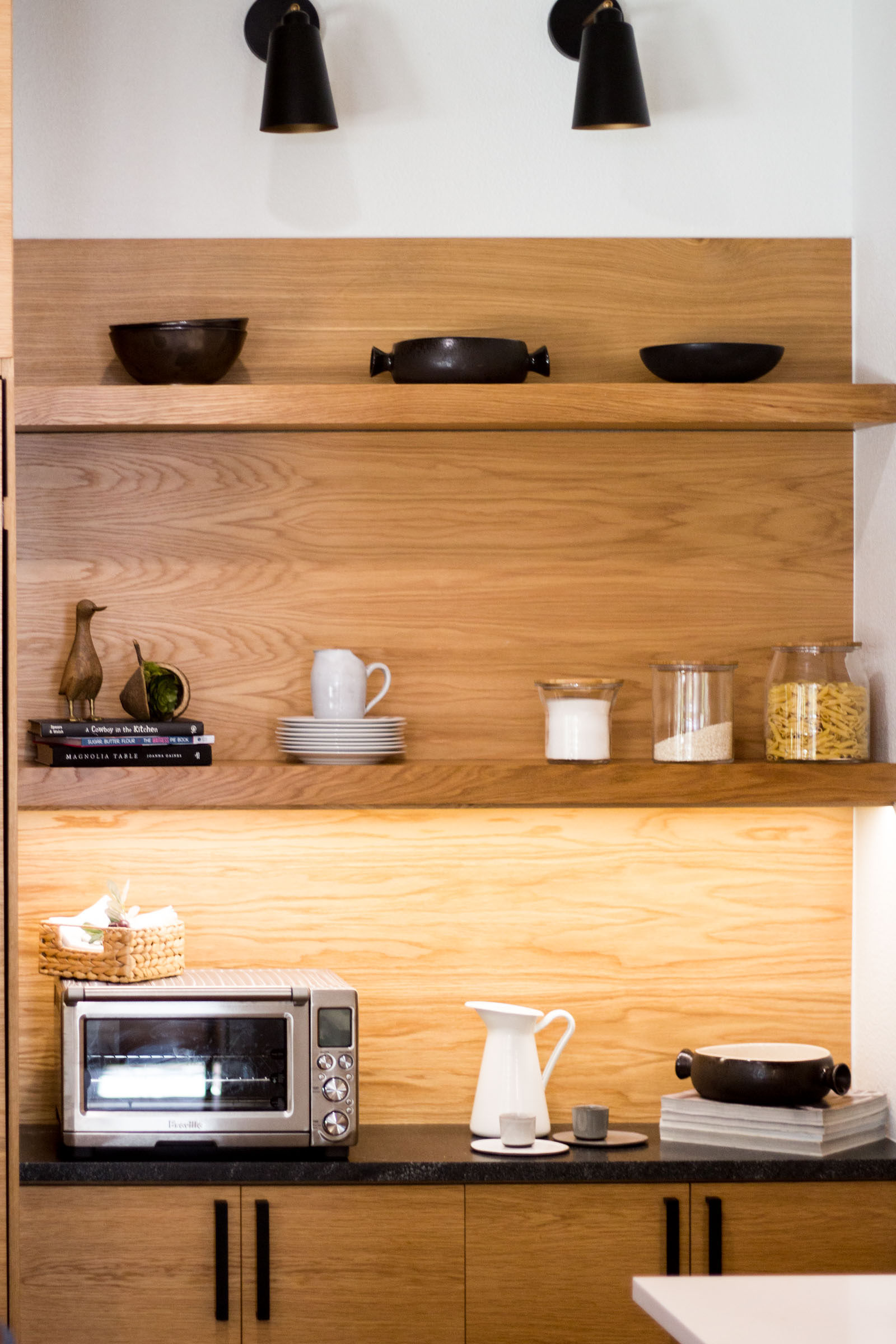 Exposed blonde wood shelves with decor and cookware, toaster oven, plates and under lighting.
