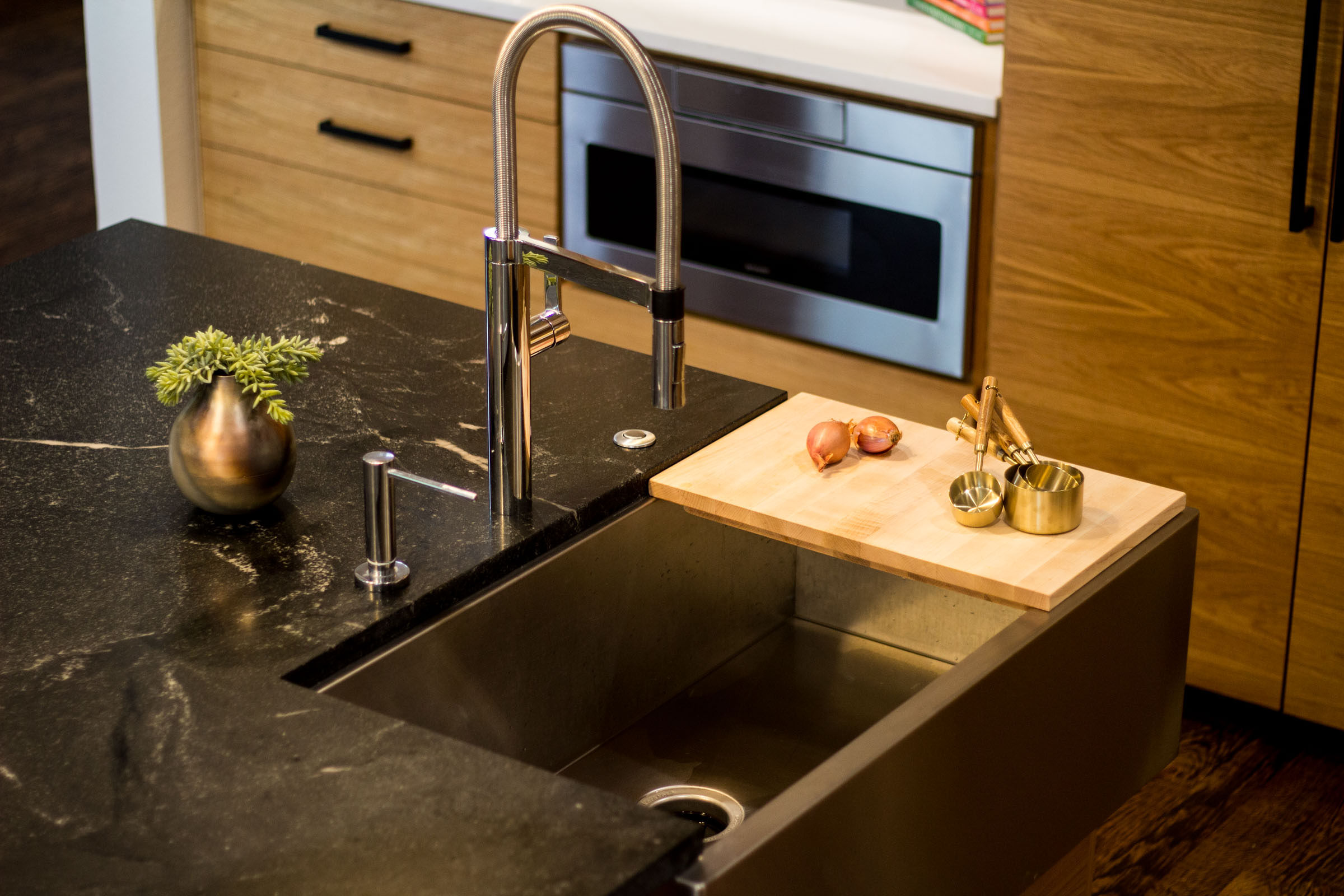 Black granite counter top, stainless steel swan kitchen faucet, cutting board onions and gold measuring cups with wooden handles.