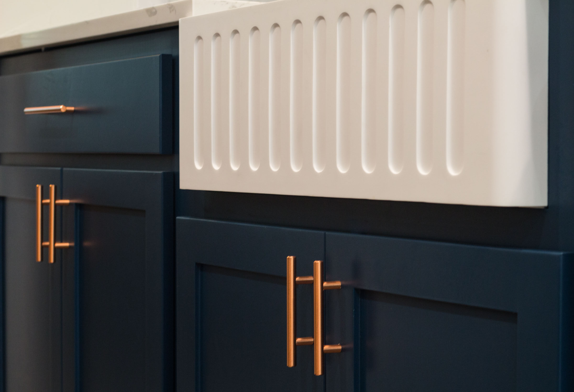 Close up of navy cabinets, copper hardware, and white farmhouse sink with grooves
